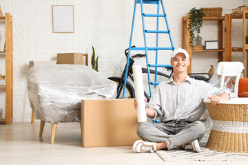 Wall Mural - Young man with roll of stretch film sitting on floor at home