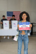 © Alan - African American woman smiles holding a 'Your Vote Matters 2024' sign at a US election polling station. In the background, a diverse group of people votes in voting booths, democracy in America