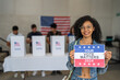 © Alan - African American woman smiles holding a 'Your Vote Matters 2024' sign at a US election polling station. In the background, a diverse group of people votes in voting booths, democracy in America