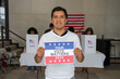© Alan - latino hispanic man holding a Your Vote Matters 2024 sign at a US election polling station. In the background, a diverse group of people votes in voting booths, democracy in America