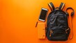 © naphat - Black Backpack with Gadgets on Orange Background: A black backpack containing gadgets such as a tablet and headphones, placed against an energetic orange background, with ample copy space on the left.