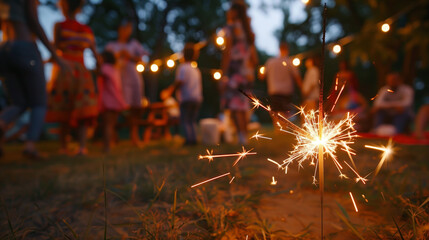 Wall Mural - A brightly lit sparkler in the foreground, casting a warm glow, with a festive backyard party in the background. 4th july, memorial. independence