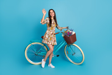 Sticker - Full body photo of attractive young woman cycling wave hand dressed stylish flowers print clothes isolated on blue color background