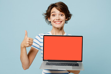 Young IT woman wears striped t-shirt casual clothes hold use work on laptop pc computer with blank screen area show thumb up isolated on plain pastel light blue background studio. Lifestyle concept.