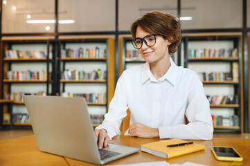 Wall Mural - Young smiling happy successful employee IT business woman wear white shirt casual clothes glasses hold use work on laptop pc computer surfing internet sit at office desk. Achievement career concept.