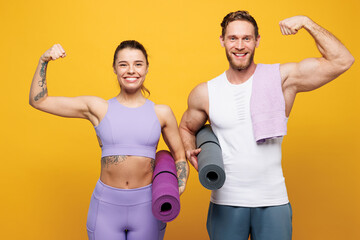 Wall Mural - Young strong fitness trainer instructor sporty two man woman wear blue clothes spend time in home gym hold yoga mat caremat show muscles isolated on plain yellow background. Workout sport fit concept.