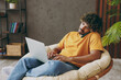 © ViDi Studio - Young IT man in orange casual clothes use work on laptop pc computer talk on mobile cell phone sits in armchair stay at home hotel flat rest relax spend free spare time in living room Lounge concept.