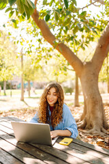 Wall Mural - Young woman sits in park with laptop. Remote work or education.  Freelance, online course.