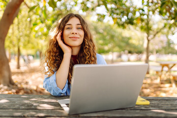 Wall Mural - Young woman sits in park with laptop. Remote work or education.  Freelance, online course.