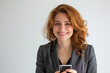 © paramee - Smiling young businesswoman with red hair holding a smartphone, wearing a dark gray blazer and black top.