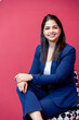 © GAJENDRRA BHATI  - Confident young indian businesswoman sitting on chair smiling looking at camera. isolated on red background. corporate and workspace concept.