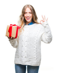 Wall Mural - Beautiful young blonde woman holding gift over isolated background doing ok sign with fingers, excellent symbol