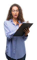 Canvas Print - Young hispanic business woman holding clipboard scared in shock with a surprise face, afraid and excited with fear expression