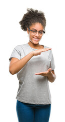 Poster - Young afro american woman wearing glasses over isolated background gesturing with hands showing big and large size sign, measure symbol. Smiling looking at the camera. Measuring concept.