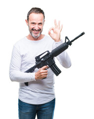 Wall Mural - Middle age senior hoary criminal man holding gun weapon over isolated background doing ok sign with fingers, excellent symbol