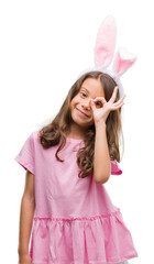 Poster - Brunette hispanic girl wearing easter rabbit ears with happy face smiling doing ok sign with hand on eye looking through fingers