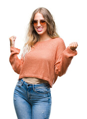 Wall Mural - Beautiful young blonde woman wearing sunglasses over isolated background very happy and excited doing winner gesture with arms raised, smiling and screaming for success. Celebration concept.