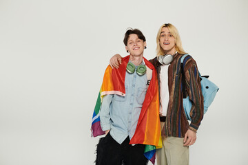 Wall Mural - Two young people stand together, smiling and holding a pride flag, on a plain white background.