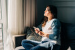 © Ilona - A woman sitting on the sofa at home by the window uses a mobile phone to scroll social networks.