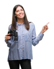 Poster - Young hispanic woman holding binoculars very happy pointing with hand and finger to the side