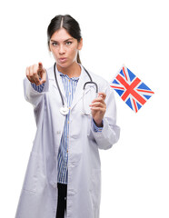 Wall Mural - Young hispanic doctor woman holding flag of united kingdom pointing with finger to the camera and to you, hand sign, positive and confident gesture from the front