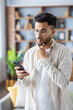© Tetiana - A young Indian man in casual and casual clothes stands at home, holds a mobile phone in his hand and stares intently at the screen, rubs his beard with his hand. Vertical photo