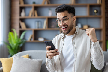 Wall Mural - Close-up photo of a happy young Nigerian man at home sitting on the sofa and happy about the news and win, looking at the phone screen and making a yes gesture with his hand