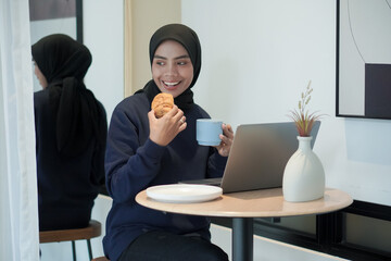 Wall Mural - A woman wearing blue sweater is sitting at a table with a laptop and a cup of coffee. She is smiling and she is enjoying her coffee and laptop