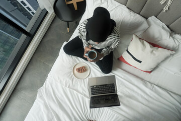 Wall Mural - A woman is sitting on a bed with a laptop and a cup of coffee. She is smiling and seems to be enjoying her time