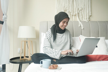 Wall Mural - A woman is sitting on a bed with a laptop and a cup of coffee. She is smiling and seems to be enjoying her time