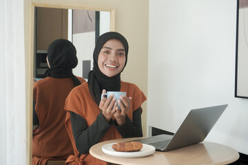 Wall Mural - A woman wearing a black scarf and a white striped shirt is sitting at a table with a laptop and a cup of coffee. She is smiling and she is enjoying her coffee and laptop