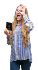 Wall Mural - Blonde teenager woman holding passport of Unites States of America with happy face smiling doing ok sign with hand on eye looking through fingers