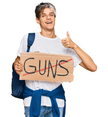 Wall Mural - Young hispanic man holding no guns warning banner smiling happy and positive, thumb up doing excellent and approval sign