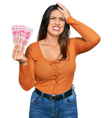 Poster - Beautiful hispanic woman holding 100 yuan chinese banknotes stressed and frustrated with hand on head, surprised and angry face