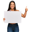 © Krakenimages.com - Beautiful hispanic woman holding blank empty banner smiling happy and positive, thumb up doing excellent and approval sign