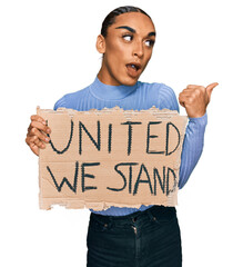 Wall Mural - Hispanic transgender man wearing make up and long hair holding united we stand banner pointing thumb up to the side smiling happy with open mouth