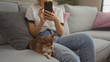 © Krakenimages.com - A young woman sits in her living room using a smartphone while her pet chihuahua rests on the couch beside her.