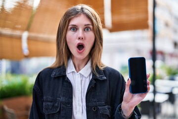 Canvas Print - Astonished young blonde woman, mouth agape in disbelief, nervously holding smartphone on bustling coffee shop terrace, embodying urban amazement