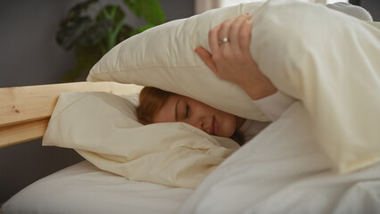 Sticker - A young caucasian woman covers her head with a pillow while lying in bed in a cozy bedroom setting.