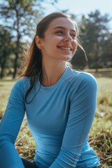 Poster - A woman is sitting in the grass wearing a blue shirt. She is smiling and looking up at the sky