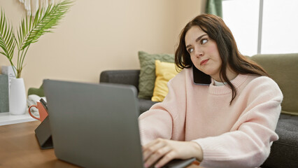 Poster - A thoughtful young woman multitasking in a cozy living room with a laptop and smartphone, exuding casual comfort and modern lifestyle.