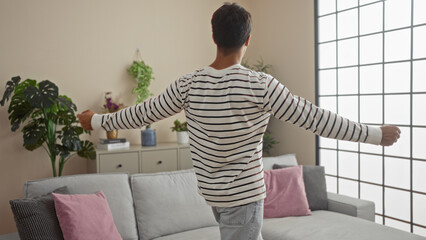 Canvas Print - A young hispanic man with outstretched arms stands in a stylish, plant-decorated living room.