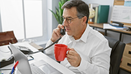 A professional middle-aged man is engaged in a conversation on the phone in a modern office setting, holding a red mug.