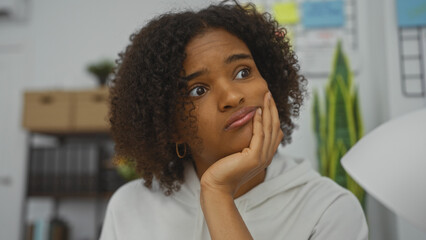 A beautiful young african american woman with curly hair is thoughtfully sitting in an office setting, showcasing a thoughtful expression in an indoor workplace environment.