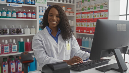 Wall Mural - A young african american woman with curly hair, wearing a lab coat, smiles while working on a computer in a brightly lit pharmacy.