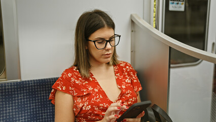 Poster - Captivating portrait of a beautiful young hispanic woman donning glasses, engrossed in her phone as she embraces urban city life, sitting alone using her smartphone on the empty subway train.