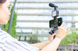 © ADDICTIVE STOCK - Young woman vlogging outdoors with smartphone and camera