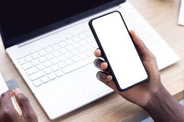 Holding smartphone with blank screen, person working on laptop at desk, copy space