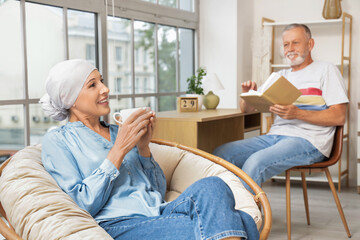 Sticker - Mature woman after chemotherapy with cup of tea sitting in armchair and her husband at home