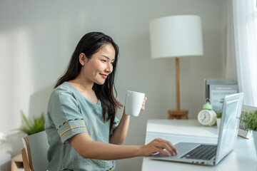 Wall Mural - Asian woman drinking coffee while using laptop in living room at home. 